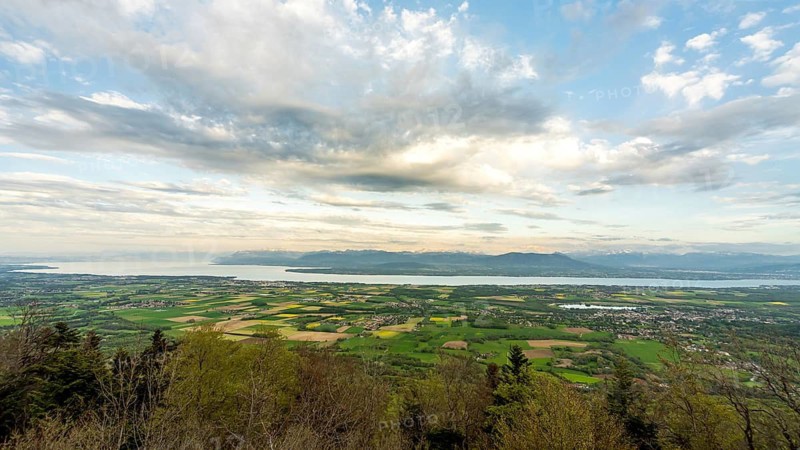 A breathtaking landscape featuring the expansive views of the Alps and Lake Leman, captured from a high vantage point near Geneva, displaying a tapestry of lush fields and cloudy skies