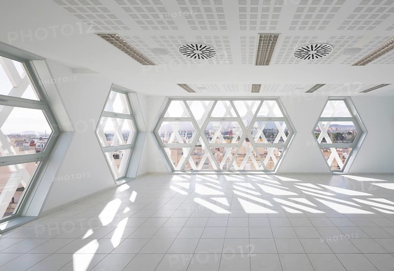 Modern auditorium interior featuring geometric, hexagonal windows. Bright natural light floods the room, creating dynamic shadows and a cityscape view