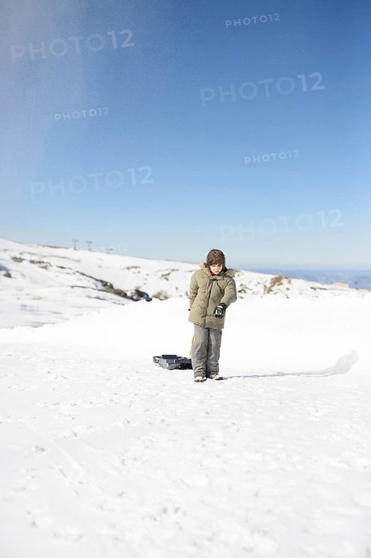 A young boy in a winter jacket stands on a snowy slope at Sierra Nevada Ski Resort, Granada, with a sled behind him. The bright blue sky and white snow create a peaceful winter scene