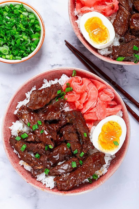 Gyudon Japanese beef with rice, pickled ginger, boiled egg slice, garnished with green onions, in a bowl with chopsticks