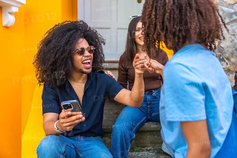 Three cheerful young friends are holding hands, laughing, and enjoying time together in front of a vibrant yellow building