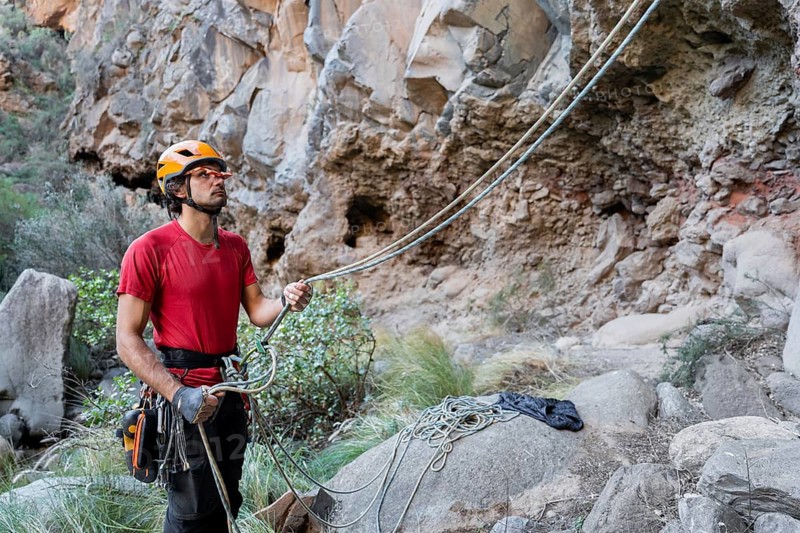 A climber in a red shirt and helmet prepares for traditional climbing on rugged rocks. The natural setting highlights the adventurous allure of outdoor mountain climbing