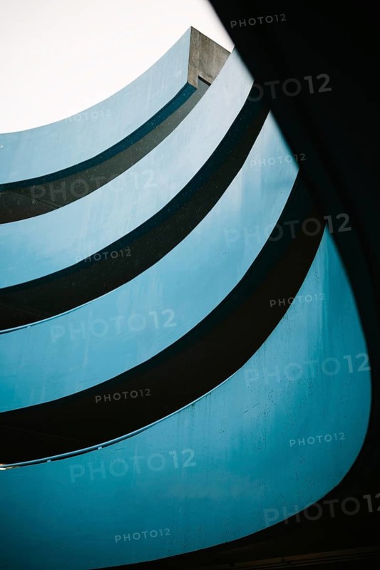 Architectural photograph of a blue parking lot building featuring multiple curved layers and a dynamic perspective. The image captures the flowing lines and vibrant blue hues of the structure, emphasizing contemporary architectural aesthetics