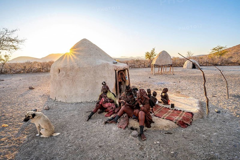 Himba woman and children sitting in front of the first woman's hut, sunrise, sun star, traditional Himba village, Kaokoveld, Kunene, Namibia