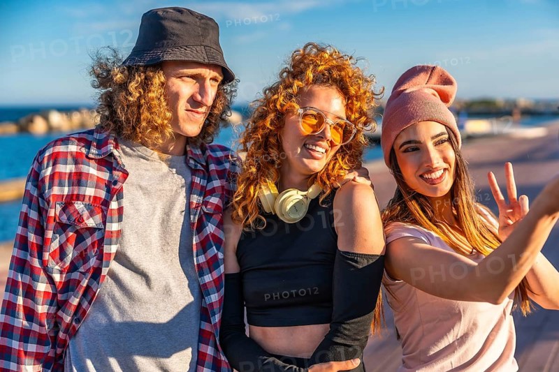 Three fashionable young friends taking a cheerful selfie while enjoying a sunny day by the seaside, radiating happiness and vibrant energy
