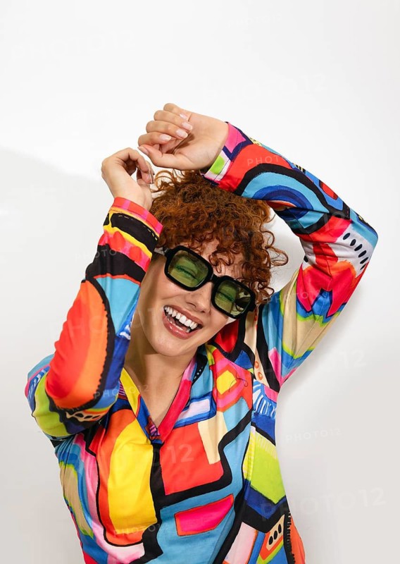 A joyful woman in a vividly patterned shirt and stylish sunglasses beams with happiness. Her playful pose and dynamic color palette exude energy and a zest for life