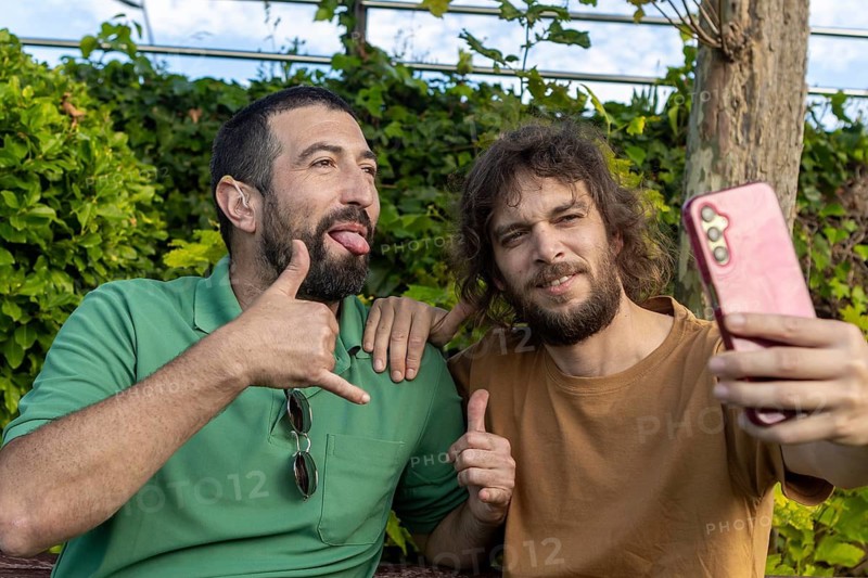 Two friends pose for a selfie outdoors in a park The man in green, who is blind, smiles and makes a playful gesture, showing joy, inclusion, and the bond of friendship in a sunny setting