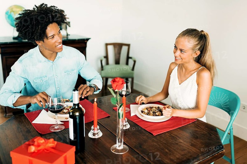 A couple enjoys a romantic dinner indoors, complete with wine, red candles, a rose, and a wrapped gift. The cozy atmosphere is perfect for a special occasion