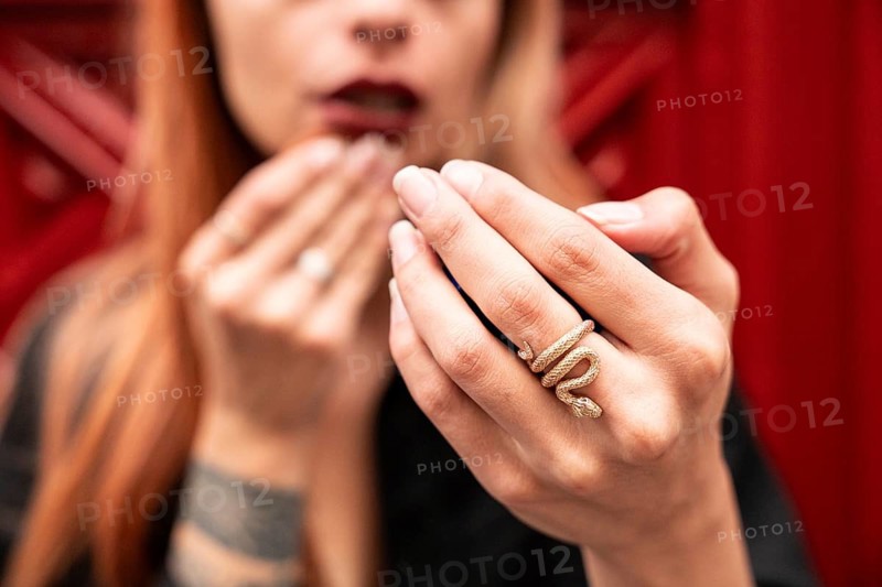 A woman with red hair shows off a gold snake ring on her finger. The background is a vibrant red, creating a striking contrast with her jewelry