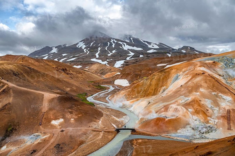 Tourist on bridge over steaming stream between colourful rhyolite mountains with snowfields, Hveradalir geothermal area, Kerlingarfjöll, Icelandic highlands, Iceland