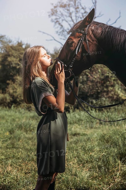 A young girl in a green dress gently interacting with a horse in a sunlit meadow, showcasing a serene bond between human and animal against a backdrop of lush trees