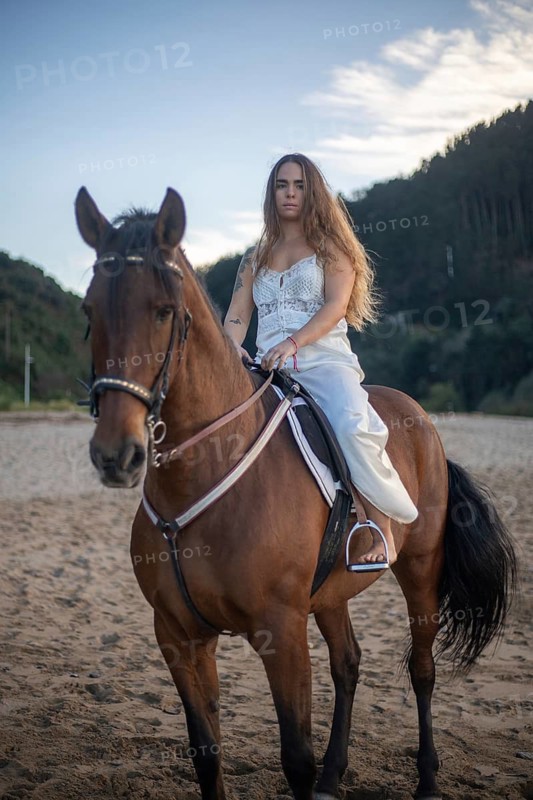A young woman with long hair rides a brown horse on a sandy beach at sunset. Dressed in white, she sits confidently, surrounded by nature and tranquility