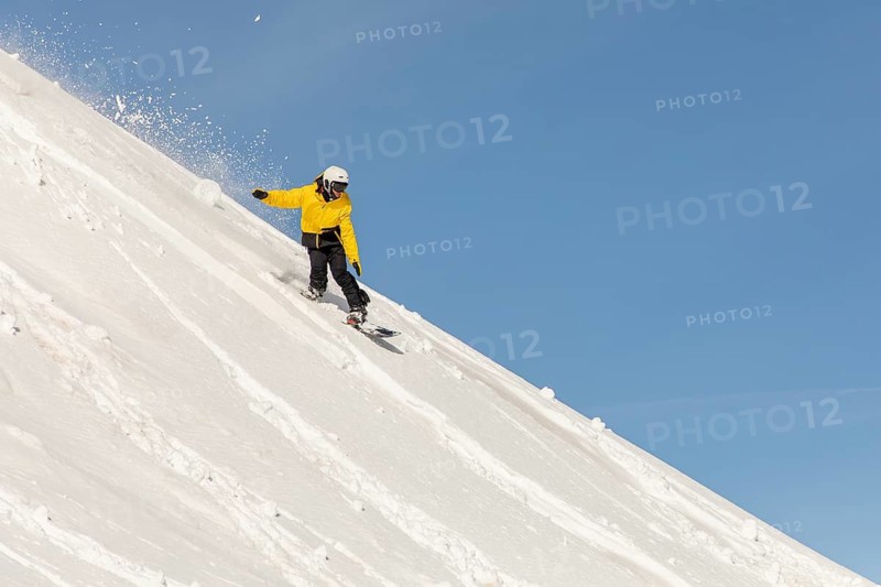 A snowboarder wearing a vibrant yellow jacket carves through fresh powder on a steep mountain slope, epitomizing the thrill of freeride snow sports in a pristine winter landscape