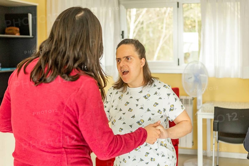 A caregiver holds hands with a resident in a mental health facility, engaging in a supportive and empathetic conversation. The setting is warm and inviting