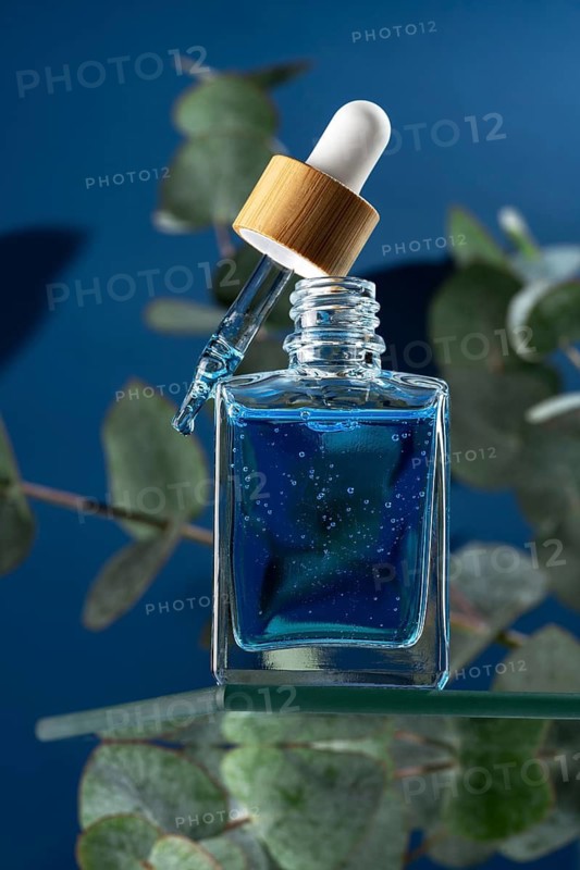A refreshing face serum designed for men, contained in a stylish glass dropper bottle with a wooden cap surrounded by eucalyptus leaves. The serum appears deep blue and features cooling properties, ideal for soothing and hydrating skin