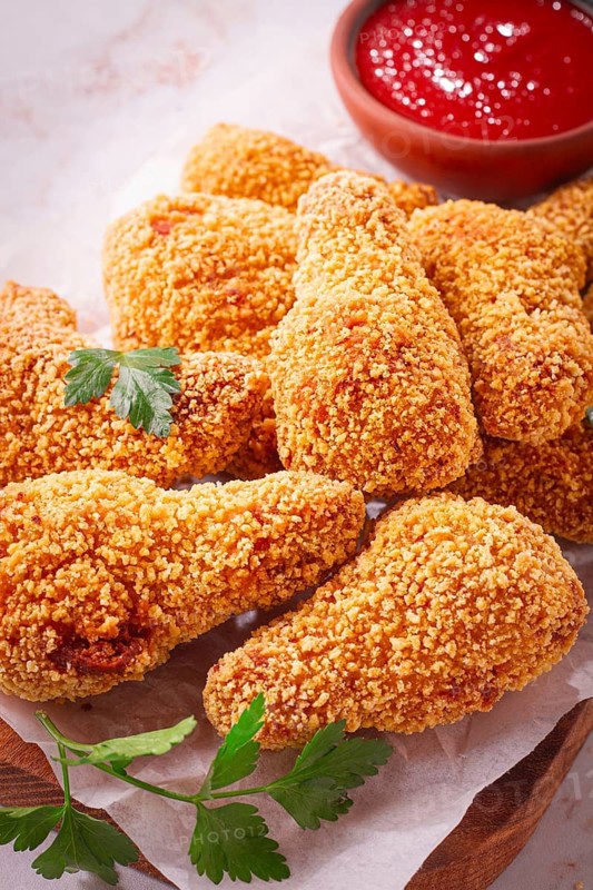A wooden board holds several golden chicken drumsticks arranged neatly. A small bowl of red dipping sauce is nearby. Fresh parsley adds a touch of color