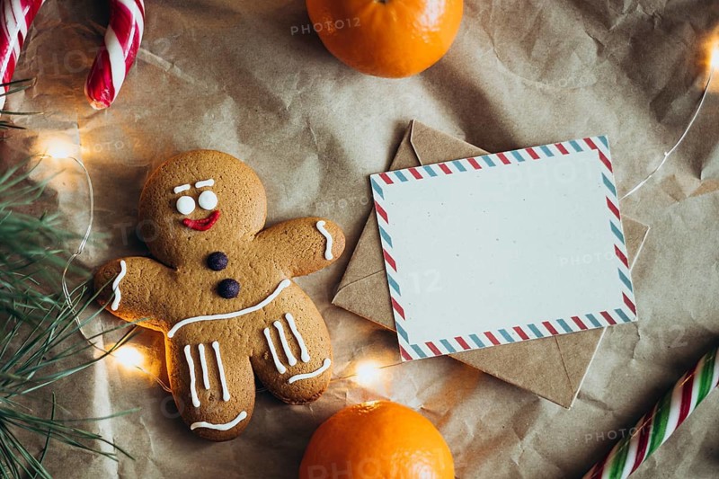 Cozy Christmas scene with a smiling gingerbread man, candy canes, a letter, oranges and fairy lights on a textured background with an empty card, perfect for festive greetings