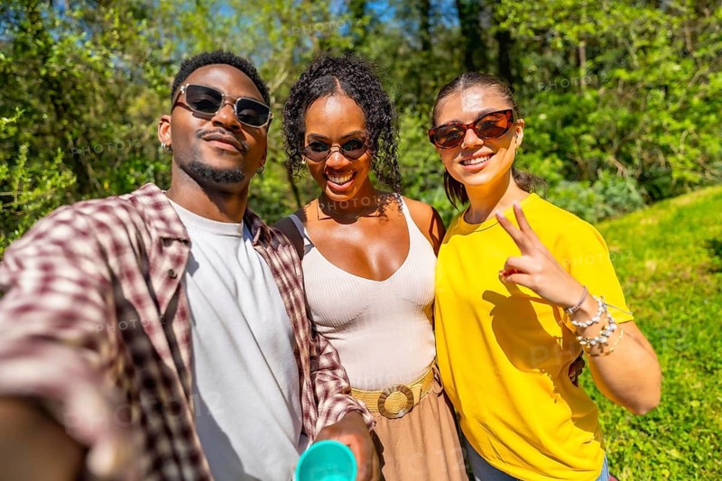 Three cheerful friends are taking a selfie in a park, enjoying their time together on a sunny day, wearing sunglasses and smiling