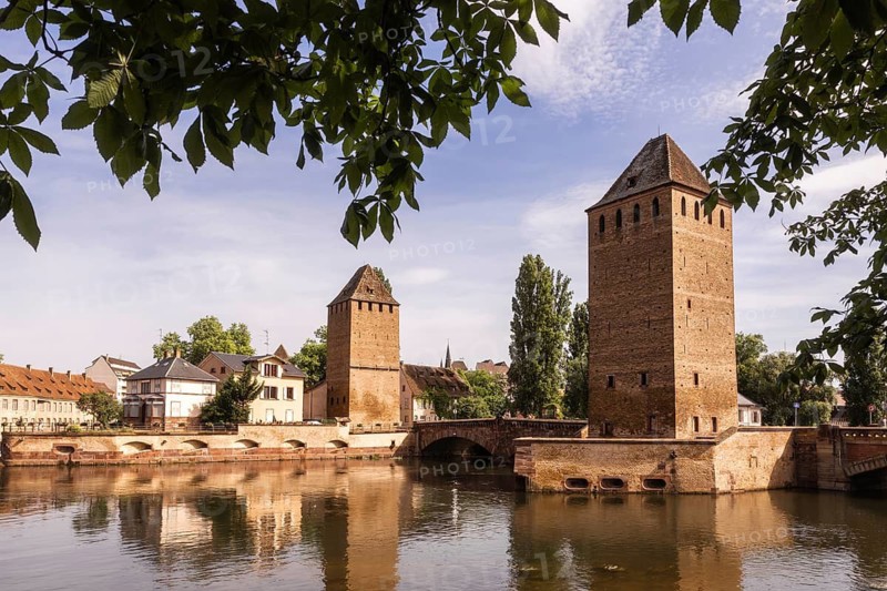 Scenic view of historic towers reflected in the calm waters of the Ill River, surrounded by lush greenery and historic architecture in the charming city of Strasbourg, France