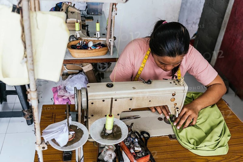 A asian seamstress focuses intently on sewing fabric using a vintage machine. The workshop is filled with tools, threads, and measuring tape, highlighting her craft and dedication