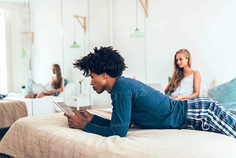 A young man lounging on a bed with a tablet, while a woman focuses on her laptop in a serene bedroom. The scene reflects a cozy, tech-savvy morning routine