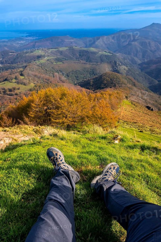 Hiker resting on grassy mountaintop, enjoying breathtaking views of rolling hills, colorful foliage, and distant ocean in autumn