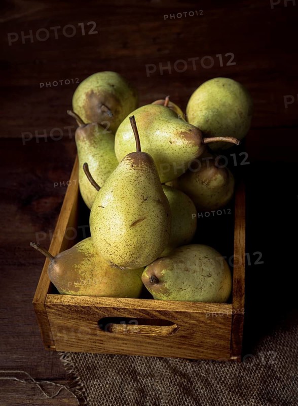 A wooden box is filled with fresh, ripe pears, resting on a burlap cloth. The dark wood background and soft lighting enhance the rustic, natural feel of the composition