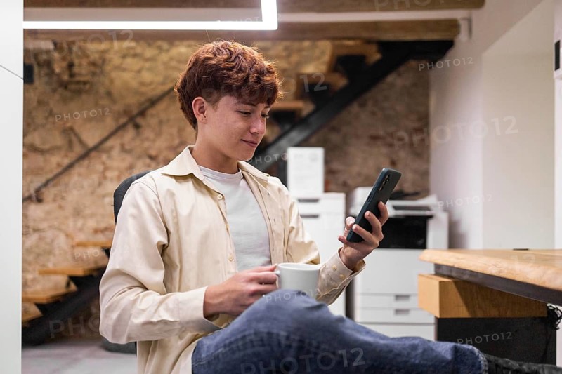 A young man in a casual shirt enjoys a summer day at a coworking space, engaging with his smartphone while holding a coffee mug, embodying a relaxed work environment