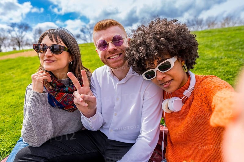 Three young diverse friends are sitting on a blanket in a park, taking a selfie and showing the peace sign