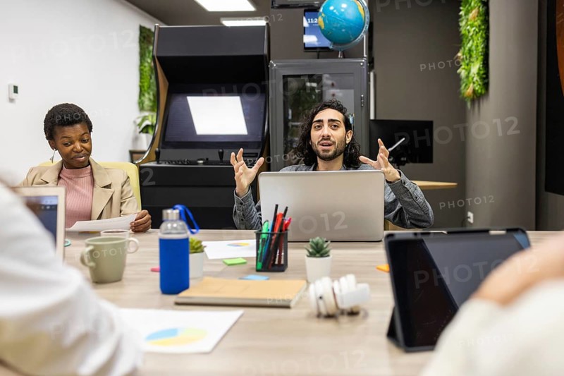 A dynamic office meeting features diverse professionals collaborating around a table with laptops, documents, and colorful office supplies in a modern, creative workspace