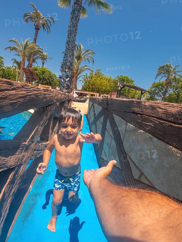 Happy child enjoying a waterslide and reaching for parent's hand at a water park, summer vacation fun