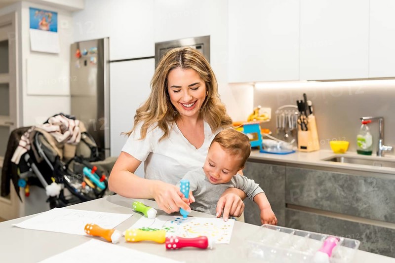 Happy mother and baby enjoying creative playtime together, using dot markers to create colorful artwork in a modern kitchen setting