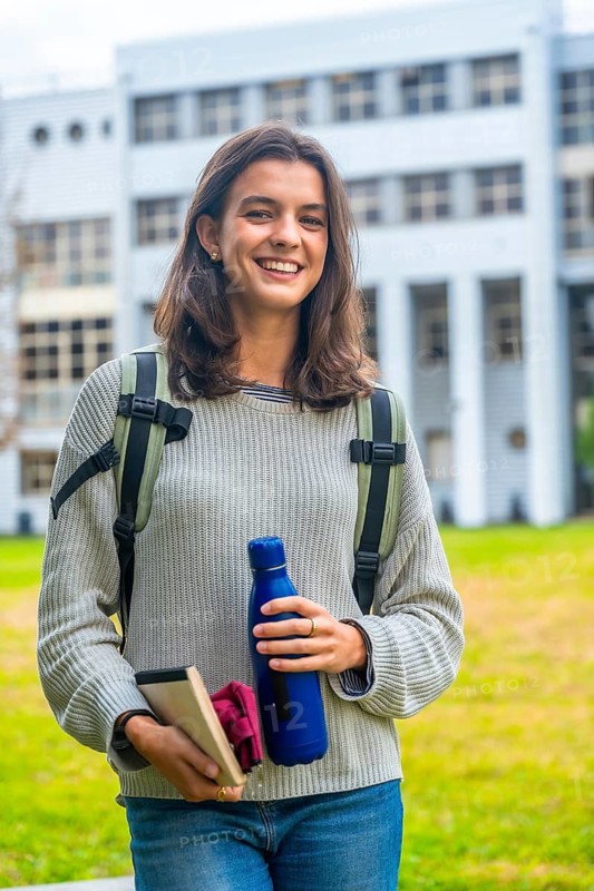 Young woman carrying a backpack and holding a reusable water bottle with books, confidently smiling outdoors on a university campus, representing education, student life, and a sustainable lifestyle