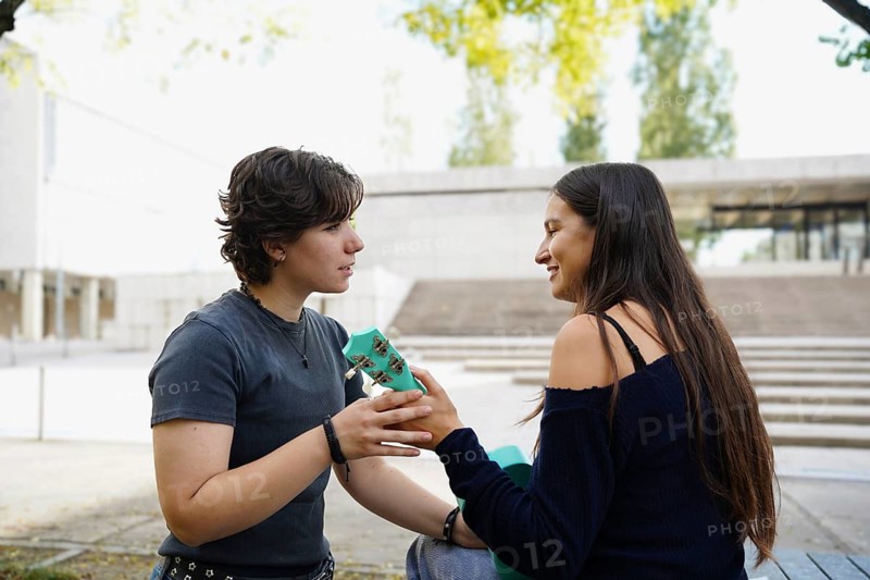 Two teenagers, a non-binary person and a female, engage in friendly conversation while seated outdoors at their school campus. They share a moment of leisure, playing the ukulele, emphasizing high school camaraderie and youth