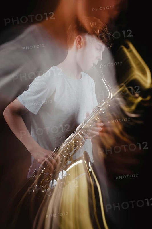 A teenage boy passionately plays a saxophone in a dimly lit studio, with dramatic lighting creating a blurred double exposure, artistic effect against a black background