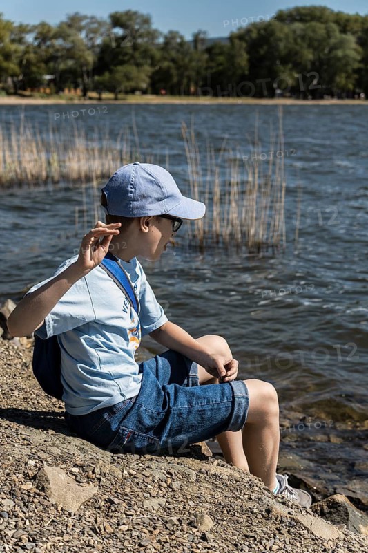 A young boy wearing a blue cap and casual attire sits by a serene lakeside, playfully engaging with nature. The sun shines brightly, creating a cheerful ambiance