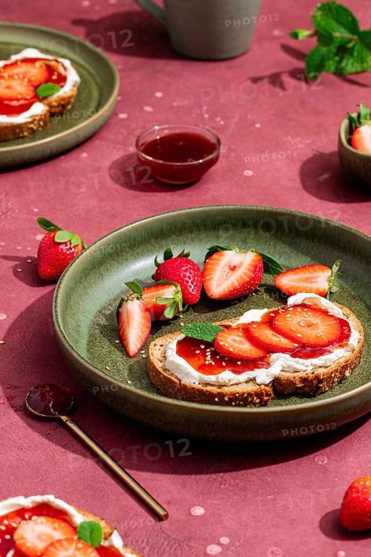 From above, a delightful brunch scene with fresh strawberries and cream cheese on toast, garnished with mint, set against a rustic red background, perfect for a summer morning