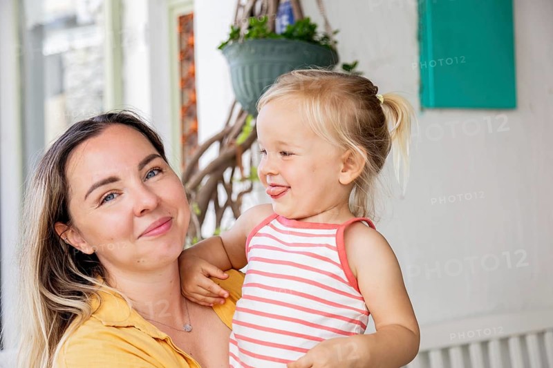A joyful mother and her young daughter share a moment outdoors. The mother is holding her daughter, who is playfully sticking out her tongue, creating a cheerful scene
