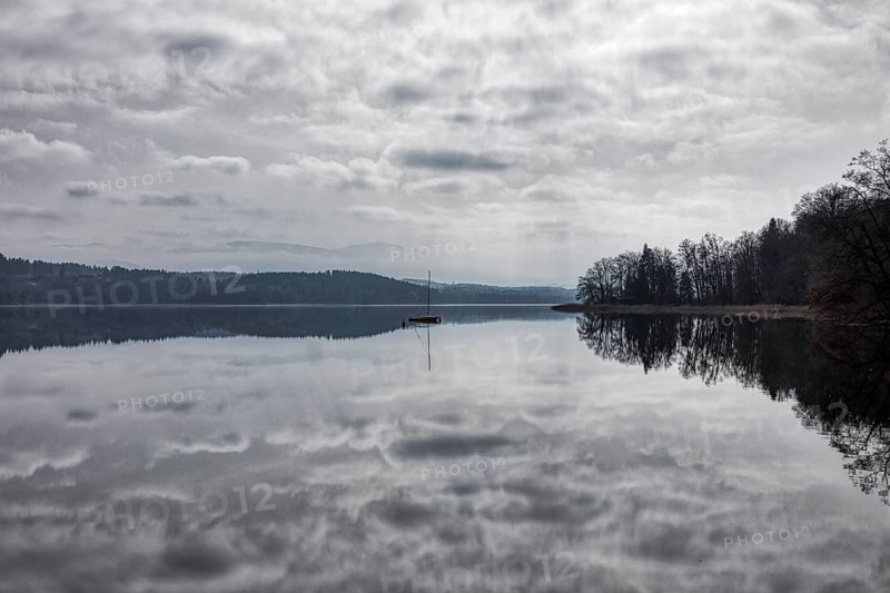 A calm lake with a single boat in the water, surrounded by forest and a cloudy atmosphere, Seehamer See, fog