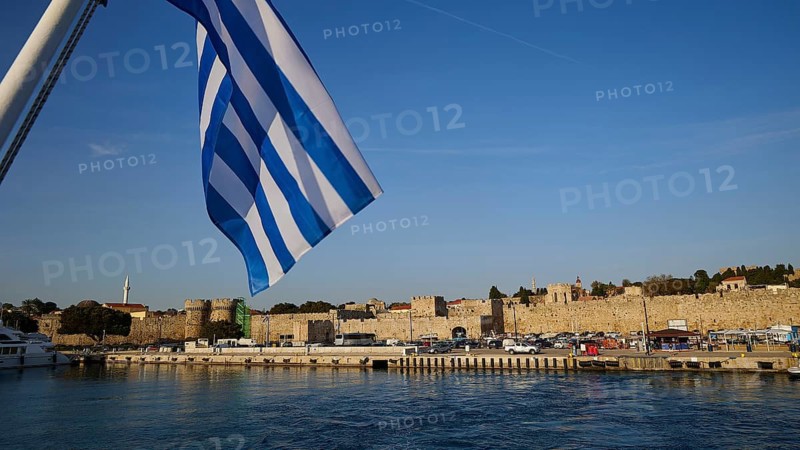 Departure of a ferry, Greek flag in front of harbour and historic city walls under blue sky, harbour area, Rhodes Town, Rhodes, Dodecanese, Greek Islands, Greece