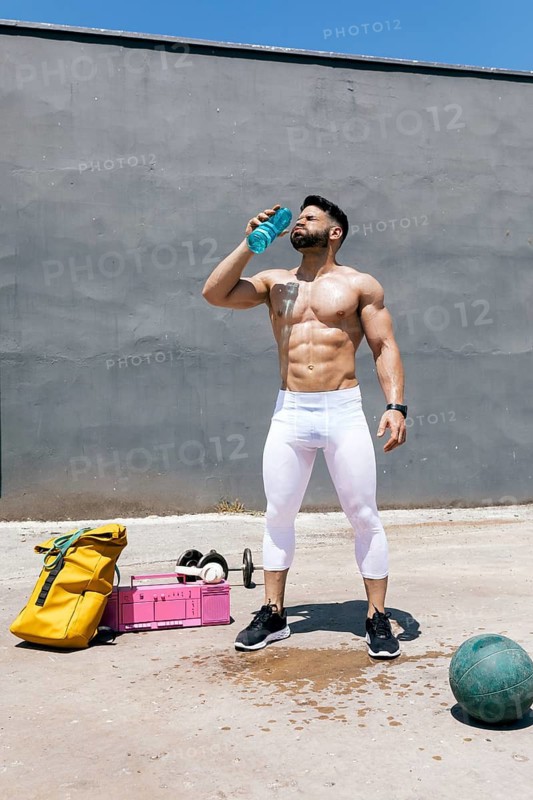 Fit man taking a break during outdoor workout, hydrating with a bottle of water under the sun. Gym equipment is scattered around, indicating an active fitness session