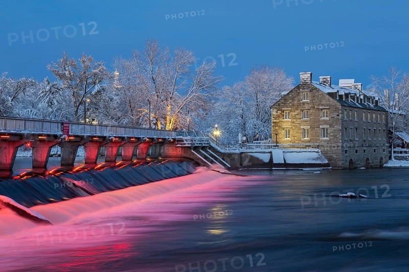 Illuminated Moulin Neuf water flow control dam and walkway over Des Mille-Iles river plus New Mill on Ile des Moulins with first winter like snow in late autumn at dusk, Old Terrebonne, Quebec, Canada