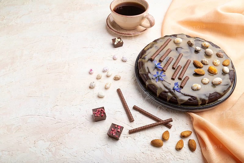 Homemade chocolate brownie cake with caramel cream and almonds with cup of coffee on a white concrete background and orange textile. Side view, close up, copy space