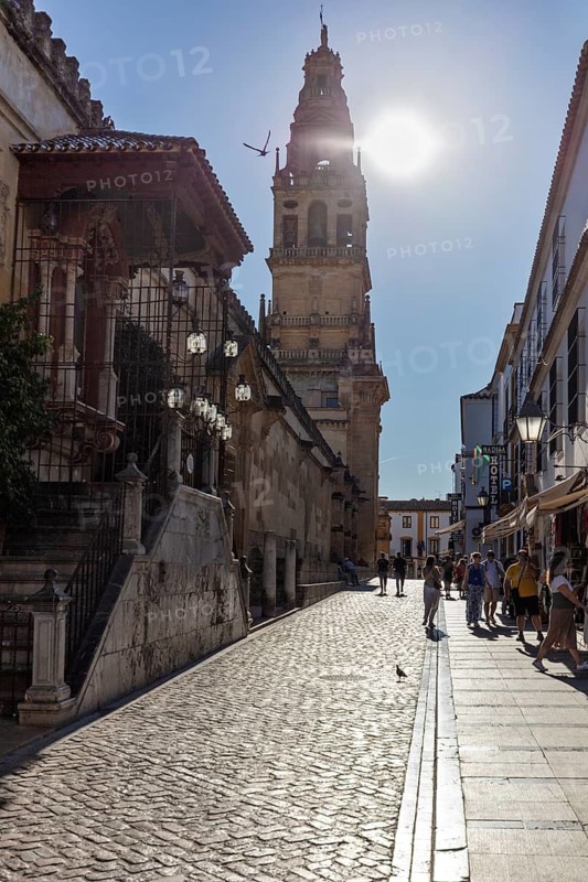 Street with church tower in the background, people walking in sunlight with long shadows, Cordoba