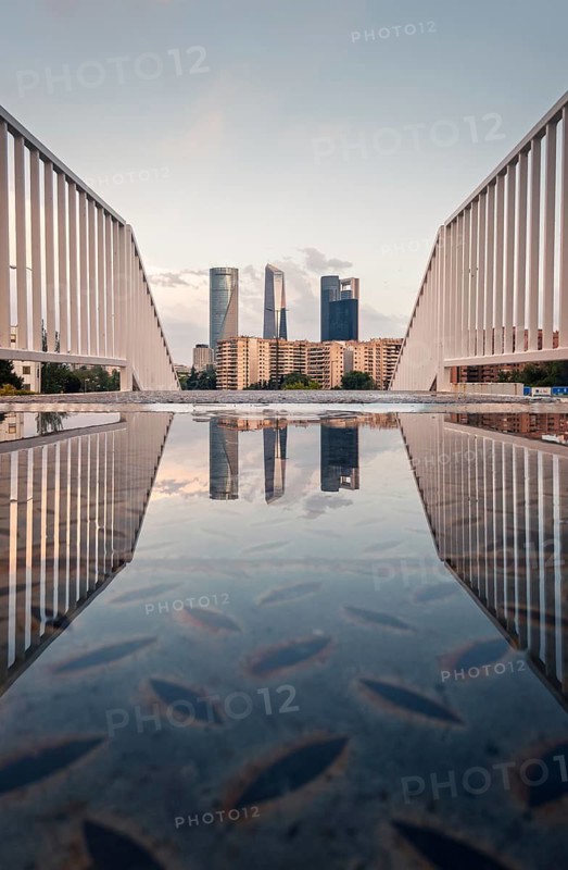 A stunning view of Madrid's skyscrapers reflected in a puddle on a bridge at sunset. The serene sky and modern architecture create a captivating urban scene