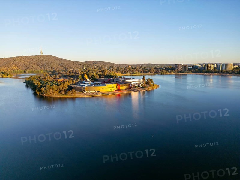 From above, this drone view of Canberra, the capital of Australia, featuring the serene waters of Lake Burley Griffin. The scene also includes a lush island hosting modern architectural structures and bordered by urban development
