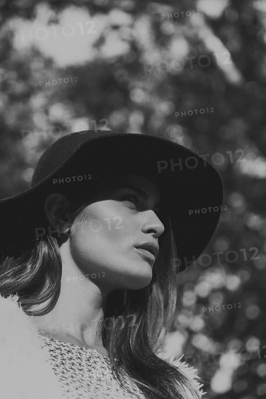 A black-and-white portrait of a woman wearing a wide-brimmed hat. Natural sunlight highlights her features against a blurred background, creating a timeless and elegant look