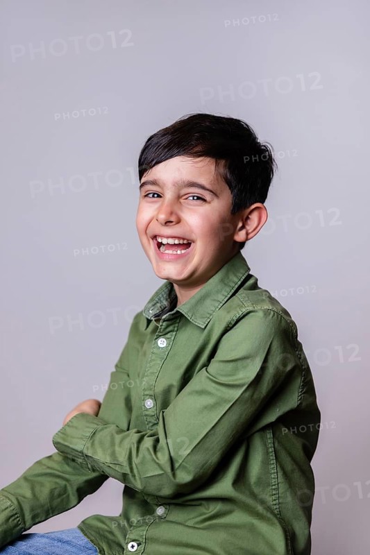 A joyful young boy with dark hair and a big smile, dressed in a green shirt and light blue jeans, sitting against a plain background in a studio setting