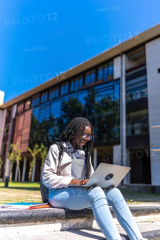 Vertical photo of a female african young student using laptop sitting outside the campus
