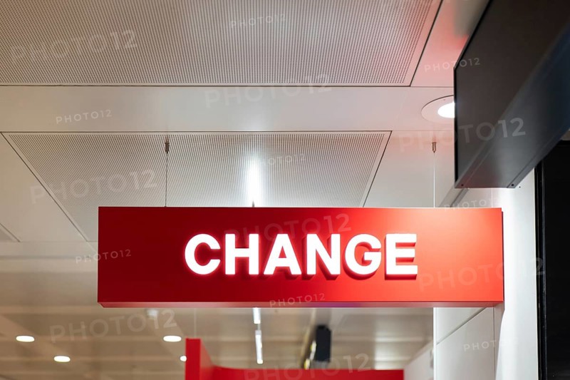 A red sign displaying the word CHANGE hangs from the ceiling of an airport terminal. The bright signage stands out against the modern interior, guiding travelers